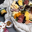 Load image into Gallery viewer, On a dark background, the local charcuterie and cheese box is plated beautifully with spreads, olives, chocolates, crackers, wine and more.