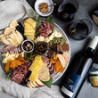 Load image into Gallery viewer, On a dark background, a silver platter of charcuterie and cheese is laid out beautifully with sine, spreads, crackers, and chocolate nearby. This photo depicts what's inside the local gift box.