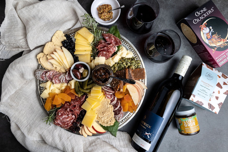 On a dark background, a silver platter of charcuterie and cheese is laid out beautifully with sine, spreads, crackers, and chocolate nearby. This photo depicts what's inside the local gift box.