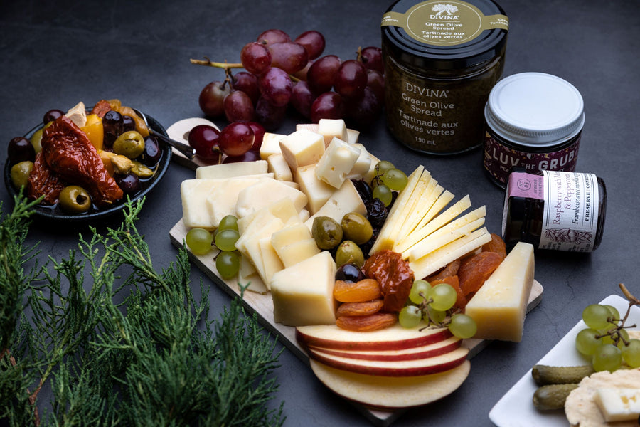 A cheese gift box is plated beautifully on a wood platter with spreads, crackers, and more laid around to depict a beautiful cheese platter.