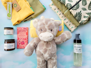 On a light blue/green background, a super cute giraffe stuffy is laying on a yellow swaddle with baby clothes, bath products, and chocolate around.