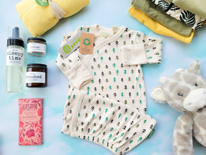 On a light blue/green background, a baby kimono with an offwhite background and greenish tree pattern is in the center, surrounded by other baby gifts.
