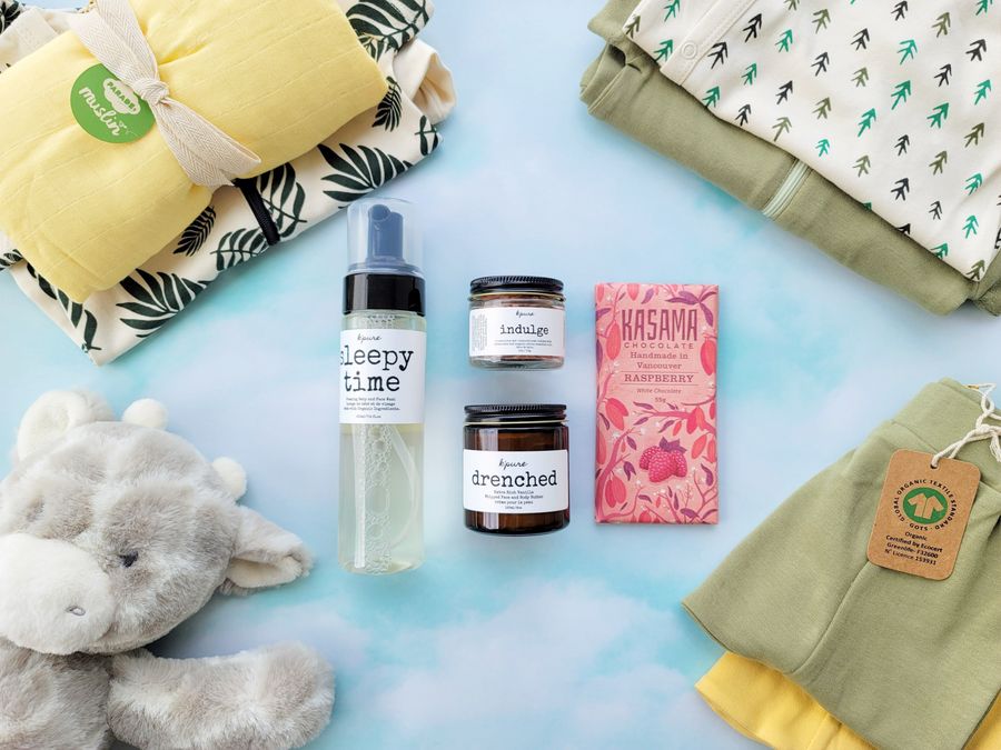 On a light blue/green background, the photo is centered on baby bath products from K'Pure and Raspberry Chocolate from Kasama, surrounded by baby clothes, swaddle, and stuffy.