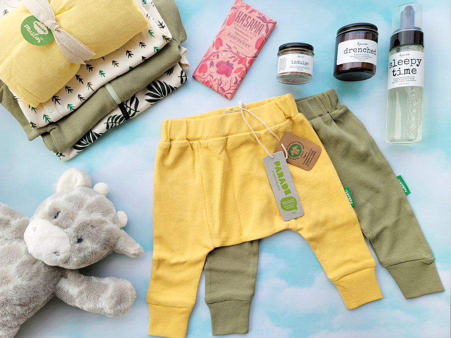 Two adorable pairs of baby harem pants in olive and light yellow are in the middle with other baby clothes, bath products, stuffy and chocolate around.