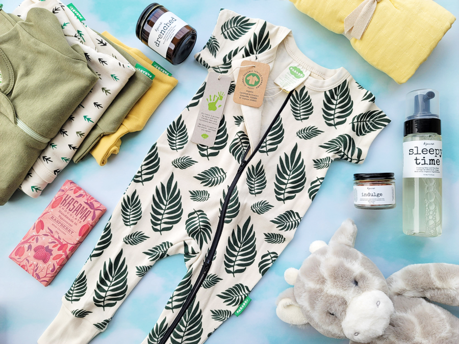 Taken from above on a light blueish green background, a very cute baby jumper from Parade is laid in the center with green ferns on an offwhite background. The jumper is surrounded by other baby gifts like a stuffie, body butter, clothing, a swaddle, and chocolate.
