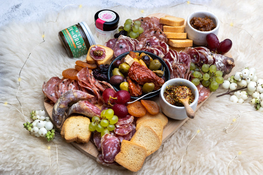 On white fur background, a beautiful charcuterie box gift is plated on a wood platter, with 3 kinds of local charcuterie, crackers, spreads, and more.