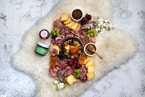 A wood platter on a white fur background is beautifully laid out a charcuterie selection with crackers, spreads, mustard, olives and more. This photo depicts the items within the gourmet charcuterie box.
