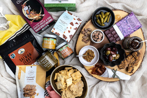On a beige linen cloth background, the contents of the hosting essentials gift box are arranged, including salmon jerky, luv the grub chutneys, kasama chocolate, almond butter ccrunch, grainy mustard, crackers, and more.