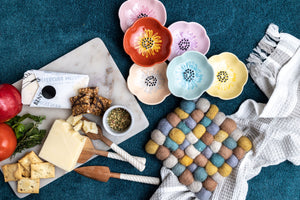 The contents of the gift basket are laid out, including the white marble board, cheese knives, multicolour trivet, towel, and six flower pinch bowls in different colours.